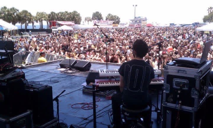 View from behind the keyboard rig facing a large daytime festival crowd under palm trees