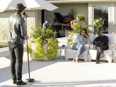 A singer performing to a mic on a sunny patio with a grand piano and two people seated, during an X Factor session
